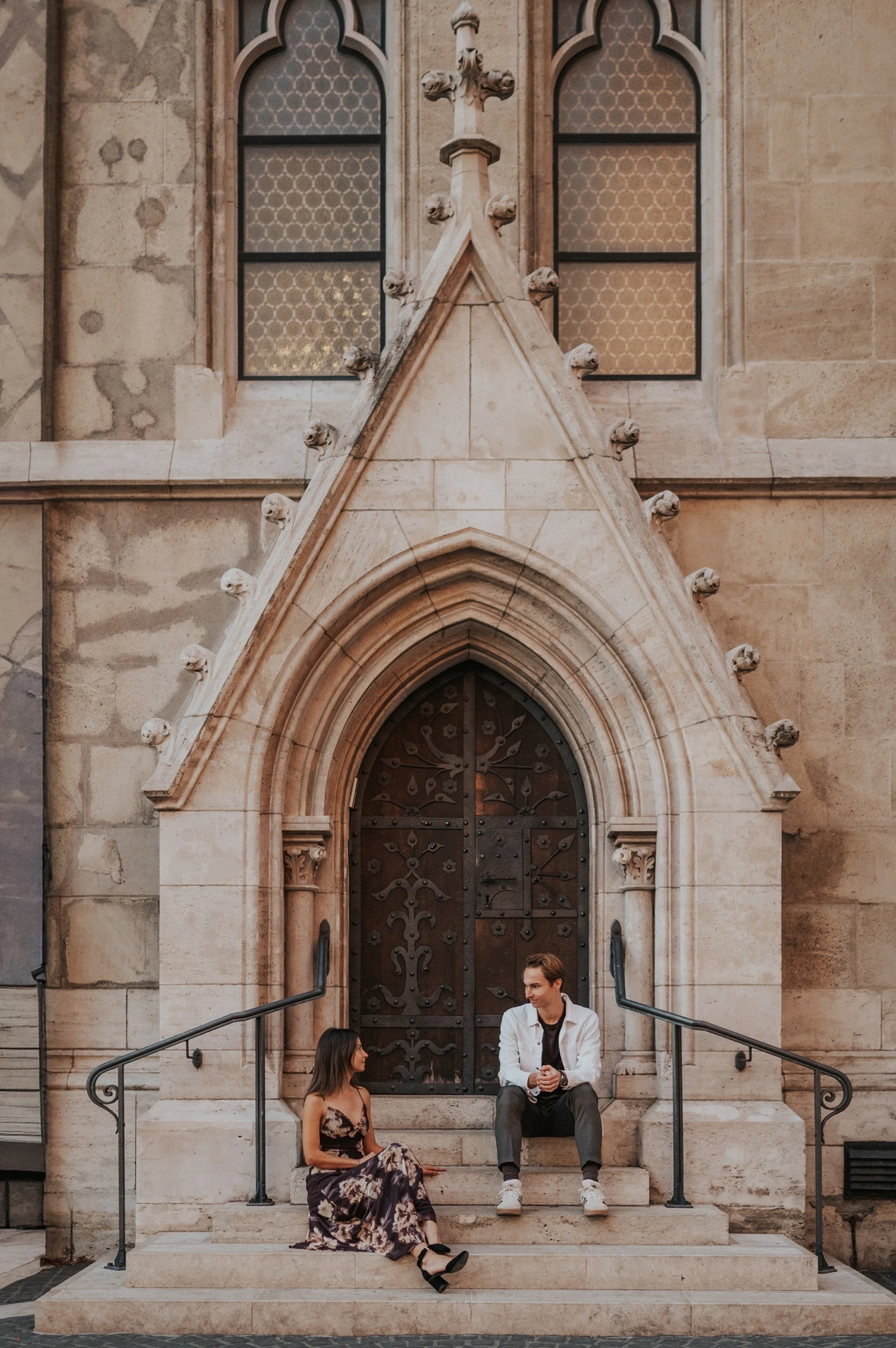 The couple sitting on opposite sides of the church entrance steps during their Budapest photoshoot, her floral dress spread across the stone.