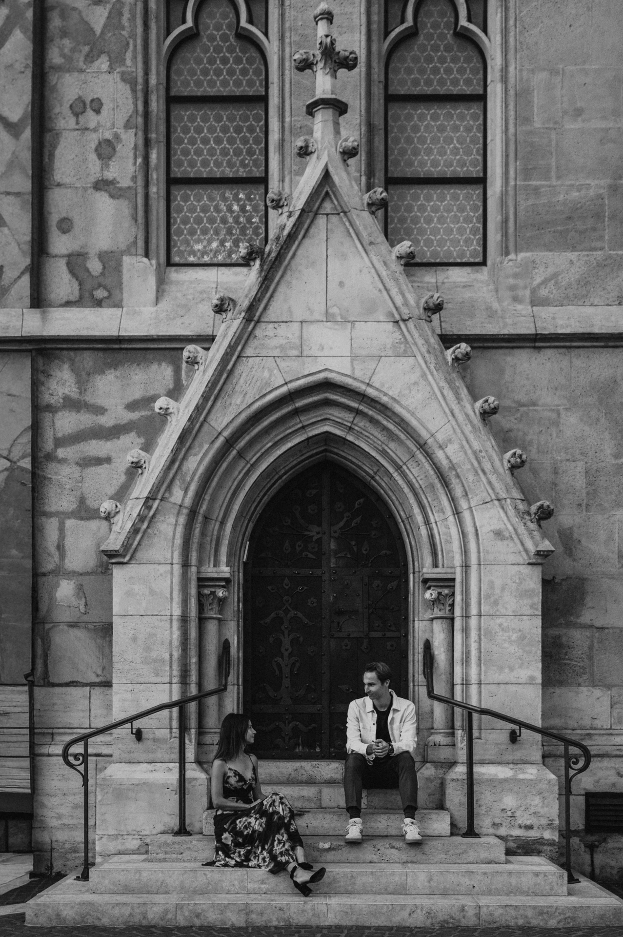 Black-and-white photograph of the couple seated beside the pointed church doorway during their Budapest photoshoot.