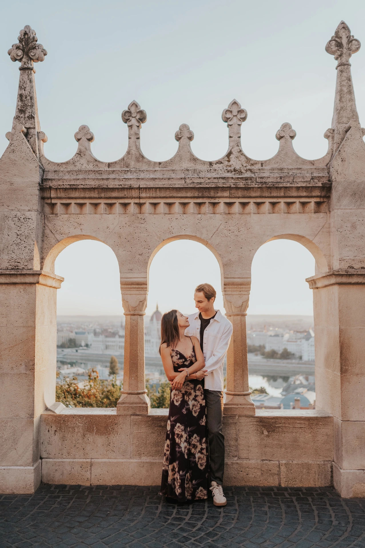 The couple embracing in front of the decorative terrace arches during their Budapest photoshoot, with the city visible beyond.