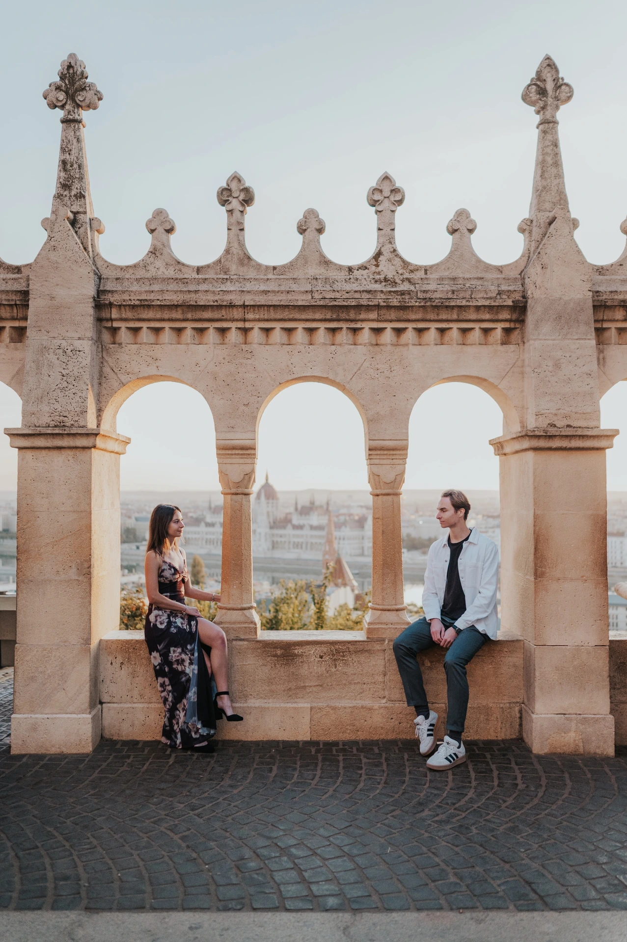 The couple seated apart along the stone terrace ledge during their Budapest photoshoot, looking towards each other between the arches.