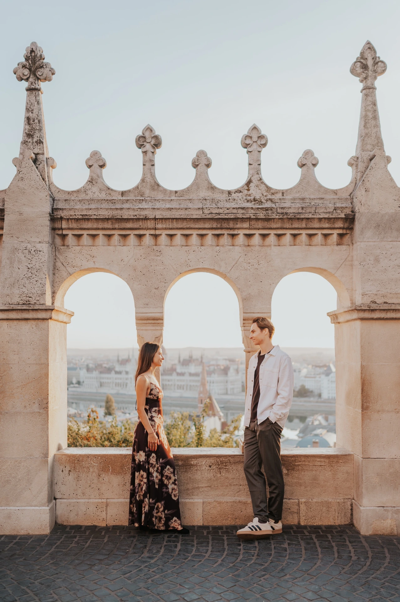 The woman seated on the stone ledge while her partner stands beside her during their Budapest photoshoot at Fisherman’s Bastion.
