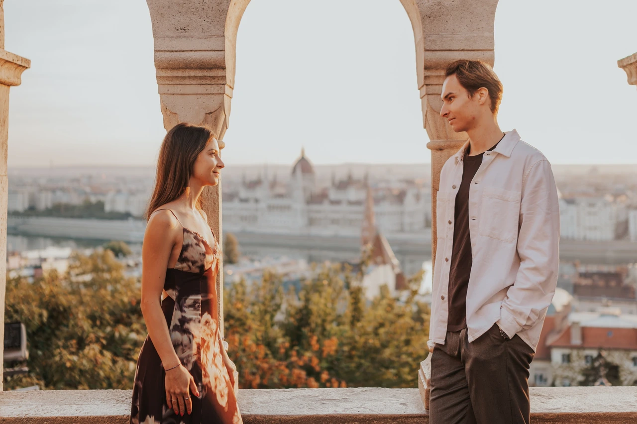 Photoshoot in Budapest with Parliament behind the terrace arches Wide portrait of the couple facing each other beneath the terrace arches during their Budapest photoshoot, with Parliament in the backdrop.