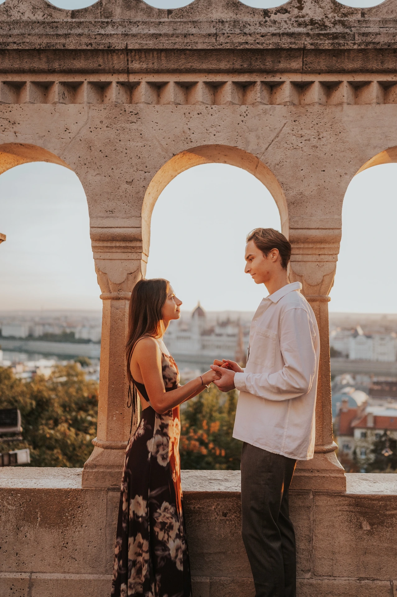 The couple holding hands in front of the open stone arches during their Budapest photoshoot, with autumn trees and the city beyond.