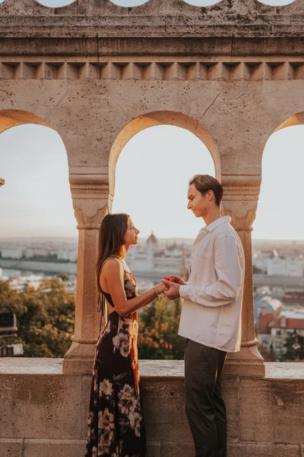 Budapest couple photoshoot holding hands at the open arches The couple holding hands in front of the open stone arches during their Budapest photoshoot, with autumn trees and the city beyond.