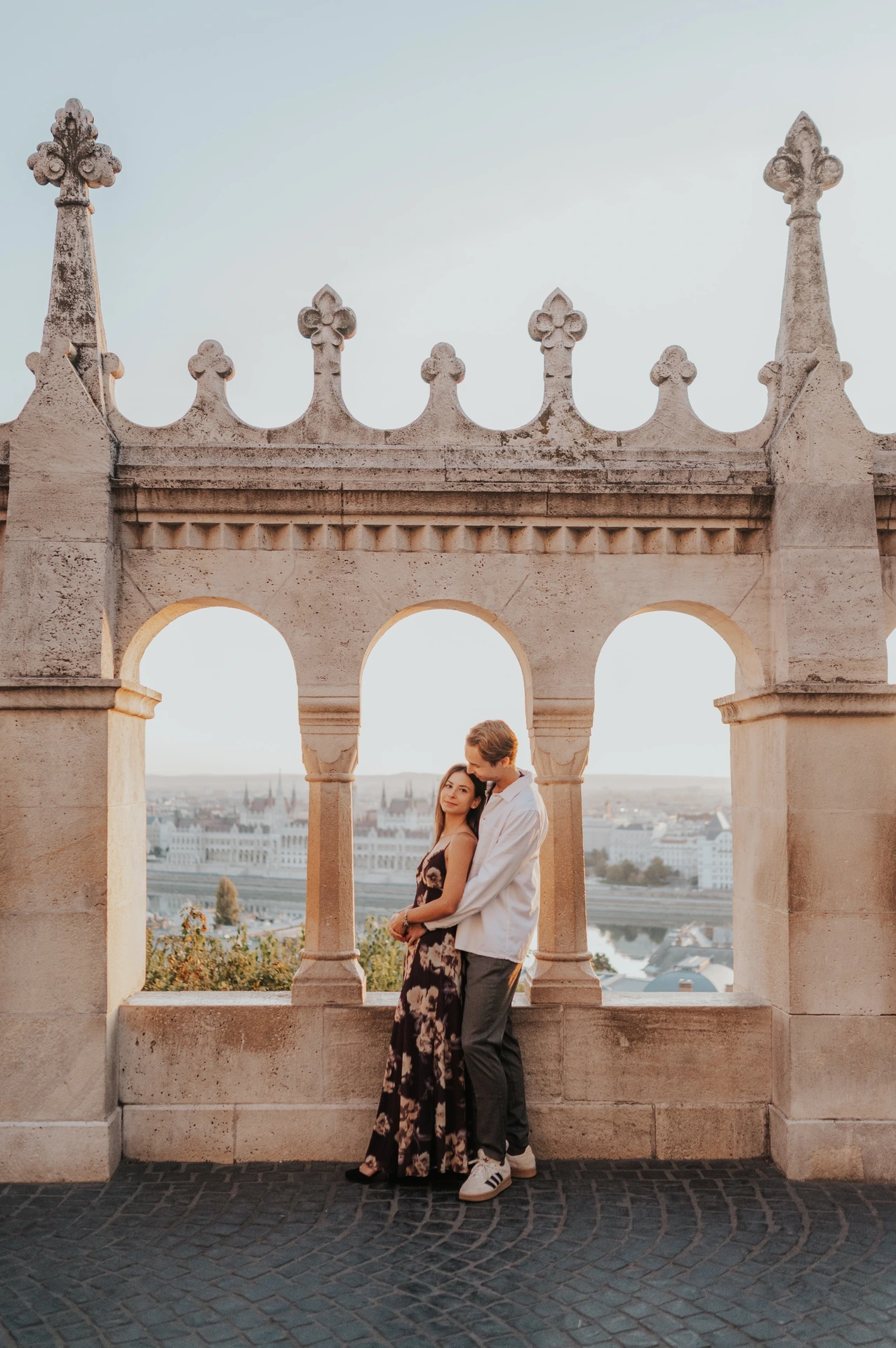 The couple leaning close together on the terrace during their Budapest photoshoot, framed by the decorative arches of Fisherman’s Bastion.