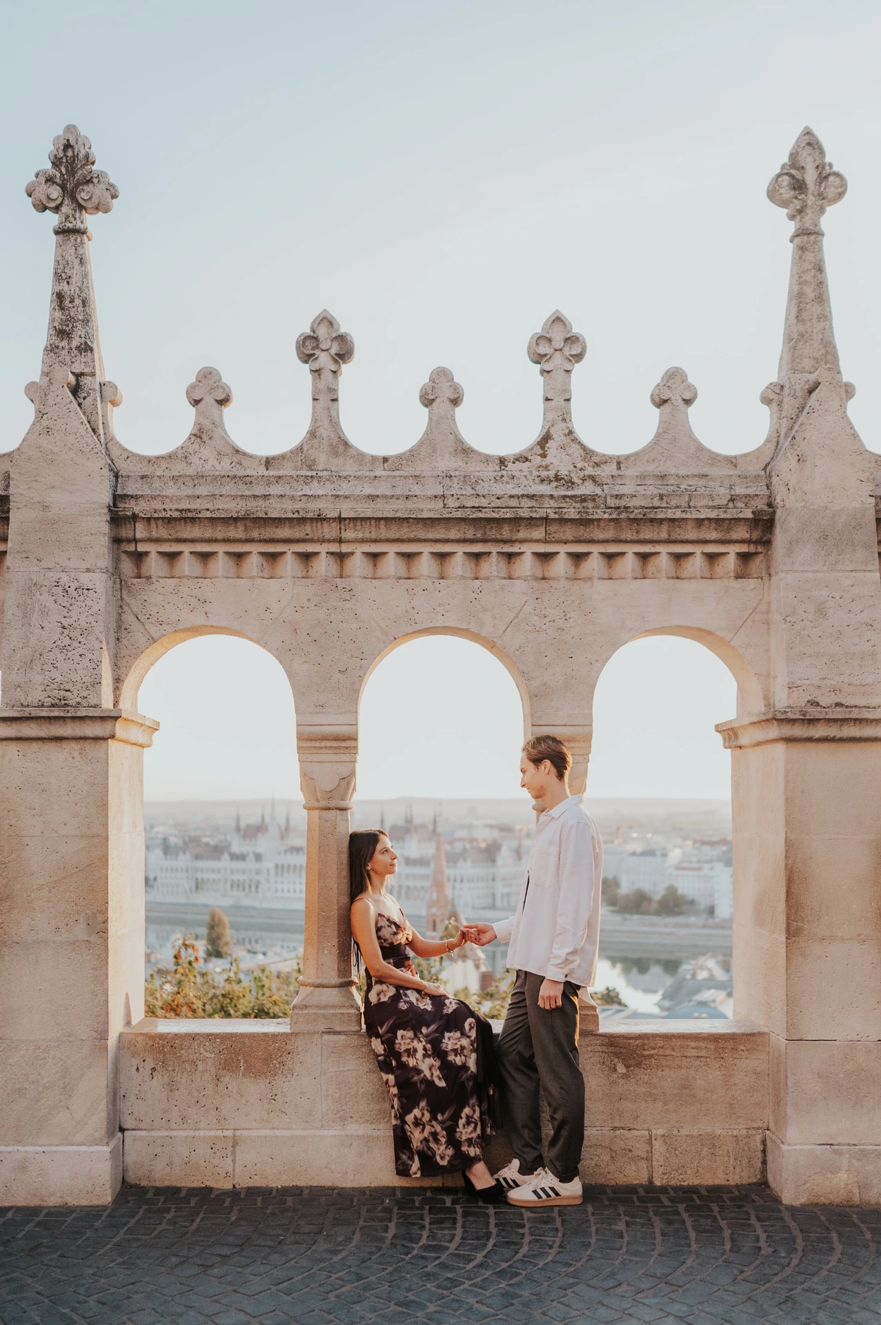 The couple sitting close together on the stone terrace ledge during their Budapest photoshoot, with the skyline visible through the arches.