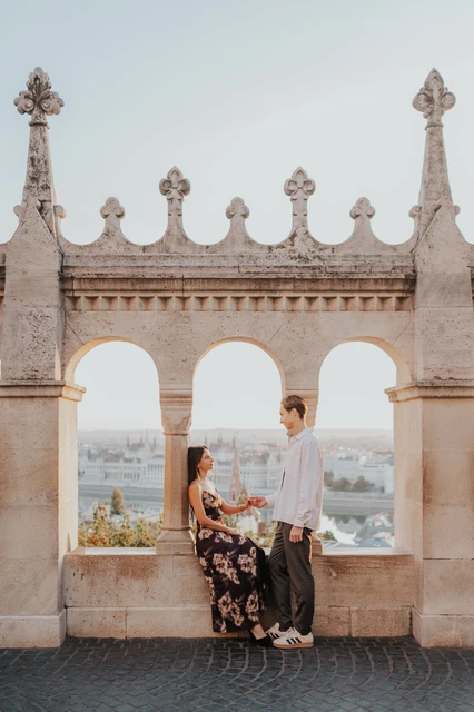 Photoshoot in Budapest sitting together beneath the terrace arches The couple sitting close together on the stone terrace ledge during their Budapest photoshoot, with the skyline visible through the arches.