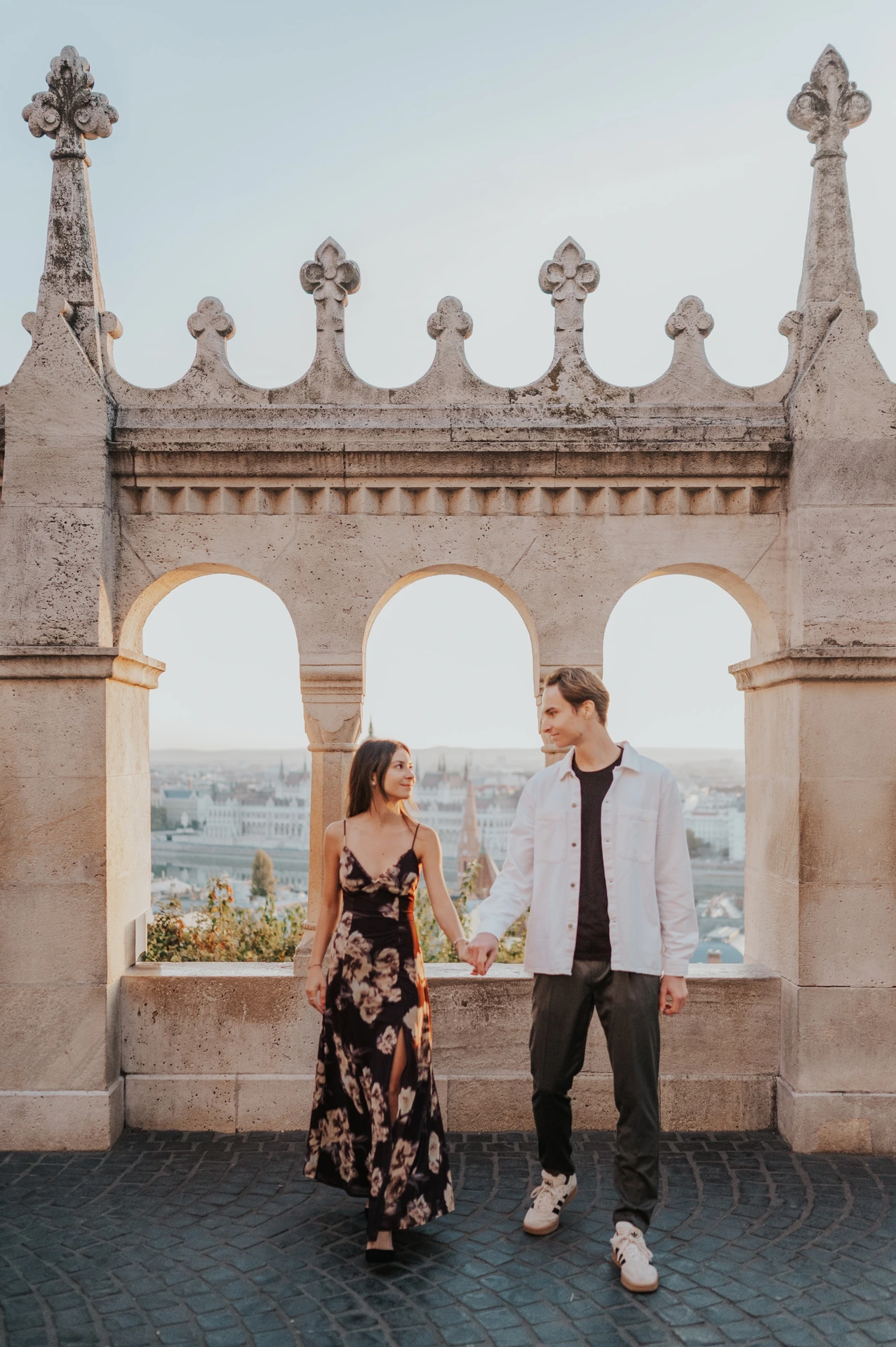 The couple standing hand in hand beneath the ornate terrace arches during their Budapest photoshoot, her floral dress catching the October light.