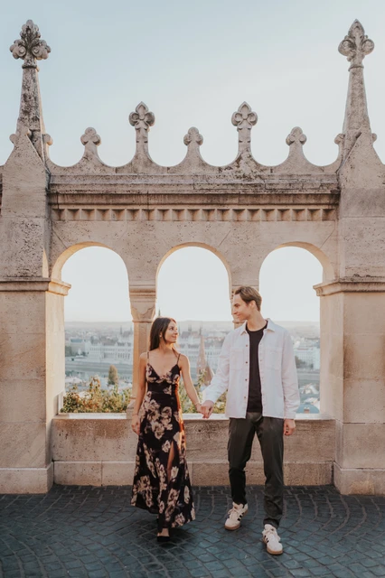 October couple photoshoot in Budapest at the ornate terrace arches The couple standing hand in hand beneath the ornate terrace arches during their Budapest photoshoot, her floral dress catching the October light.