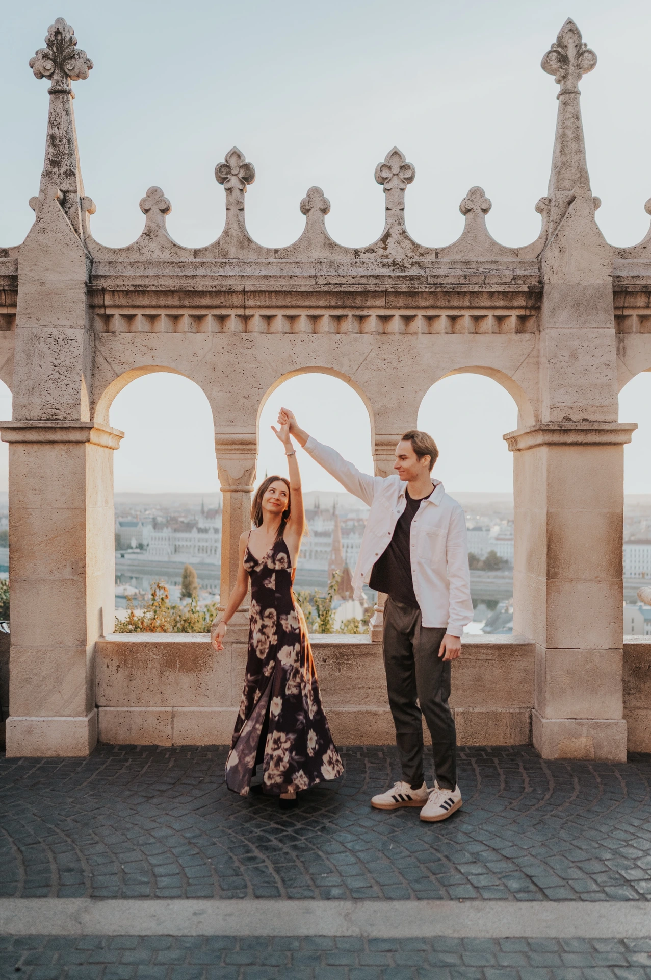 The man lifting their joined hands as his partner twirls in her floral dress during their Budapest photoshoot at Fisherman’s Bastion.