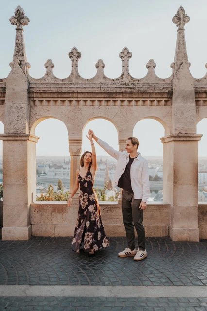 The man lifting their joined hands as his partner twirls in her floral dress during their Budapest photoshoot at Fisherman’s Bastion.