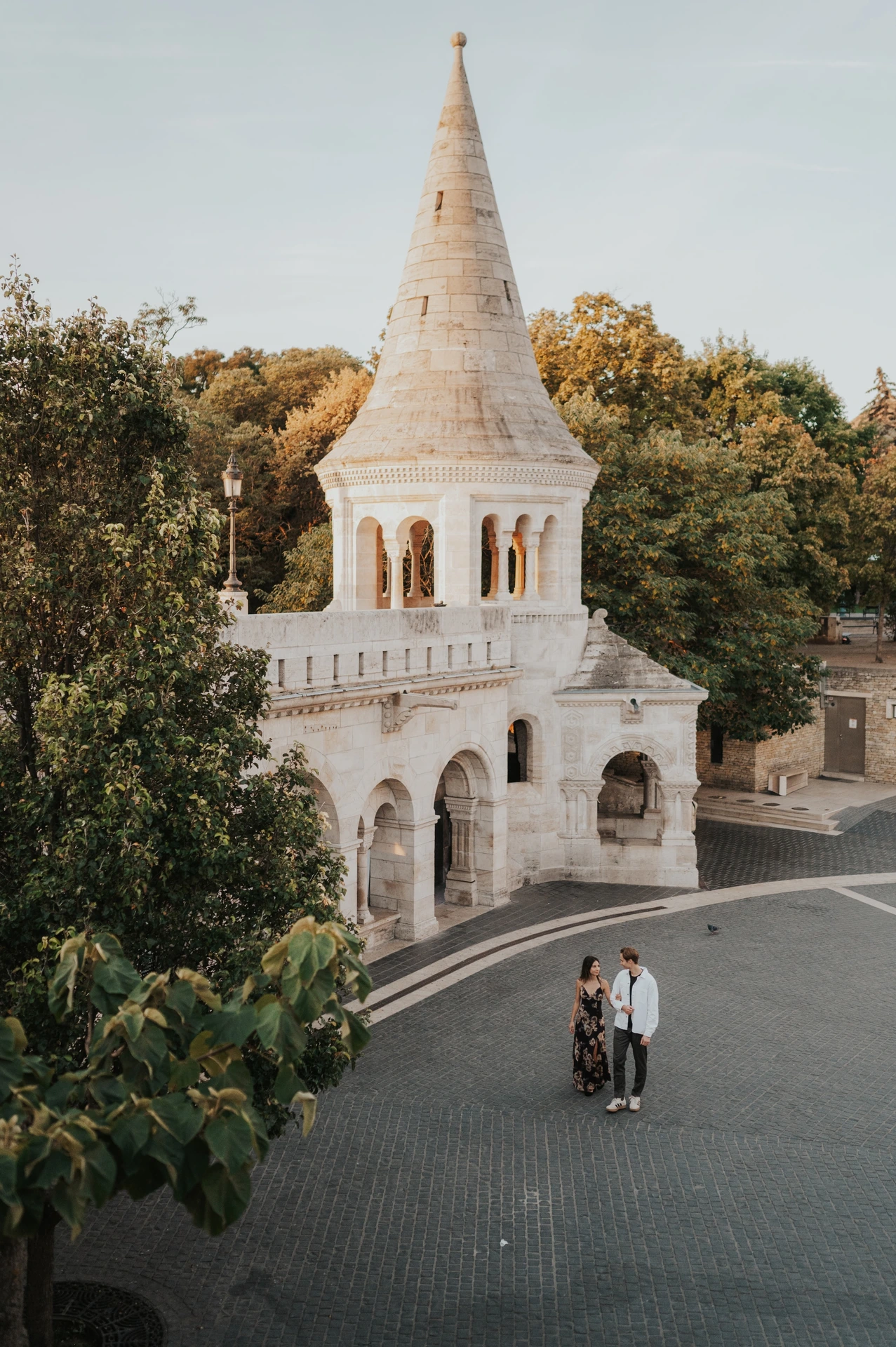 The couple standing together below a stone turret with autumn trees behind it during their Budapest photoshoot at Fisherman’s Bastion.