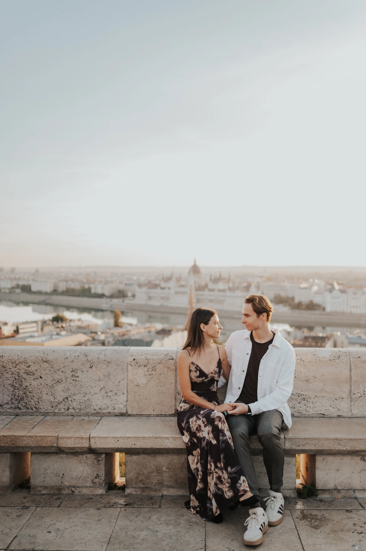 The couple sitting on a stone bench overlooking the city during their Budapest photoshoot, with a wide blue sky above them.