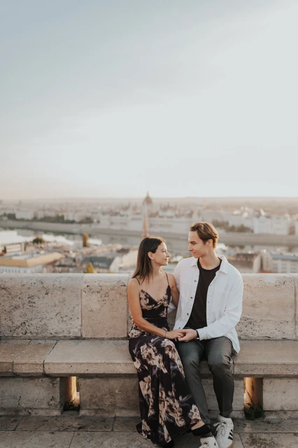 Photoshoot in Budapest with Parliament behind the seated couple The couple smiling towards each other on a stone bench during their Budapest photoshoot, with Parliament across the city behind them.