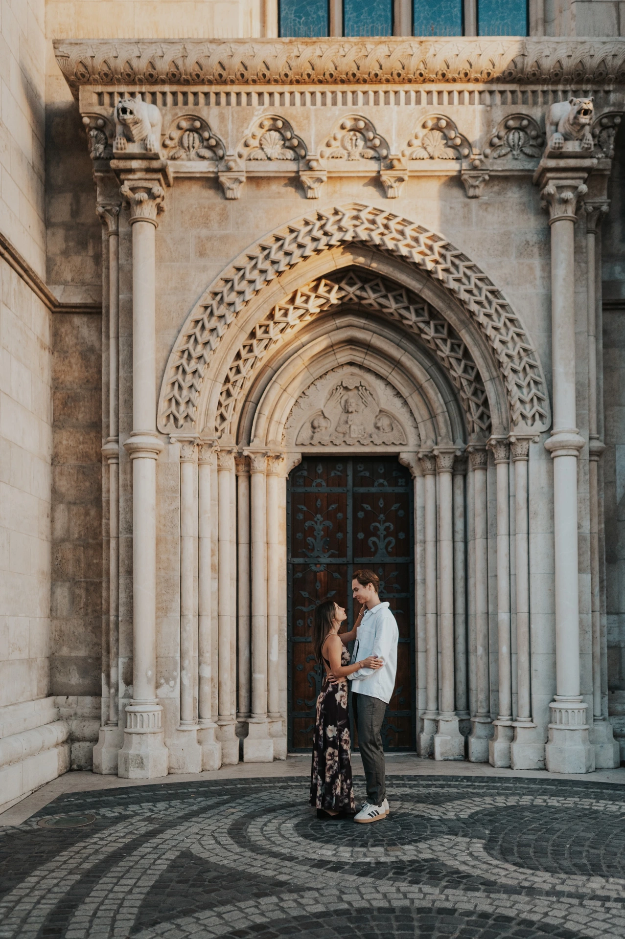 The couple embracing below the richly carved church arch during their Budapest photoshoot, surrounded by stone columns and patterned paving.