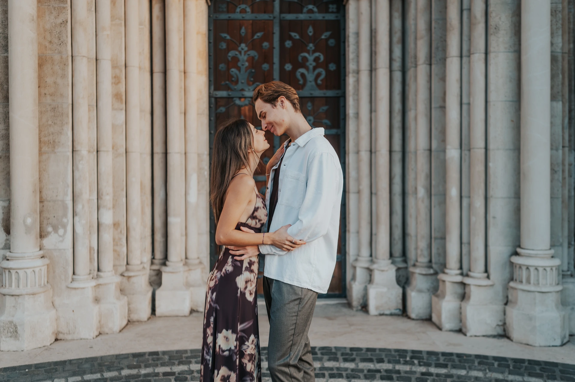 Budapest couple photoshoot embracing at the carved church doorway The couple holding each other close in front of the carved church doorway during their Budapest photoshoot, her floral dress against the pale columns.