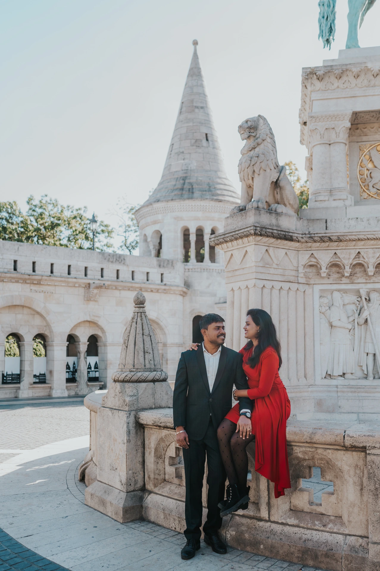 A wide portrait of the couple beside a carved monument with Fisherman’s Bastion’s pointed turret and arches behind them during their Budapest photoshoot.