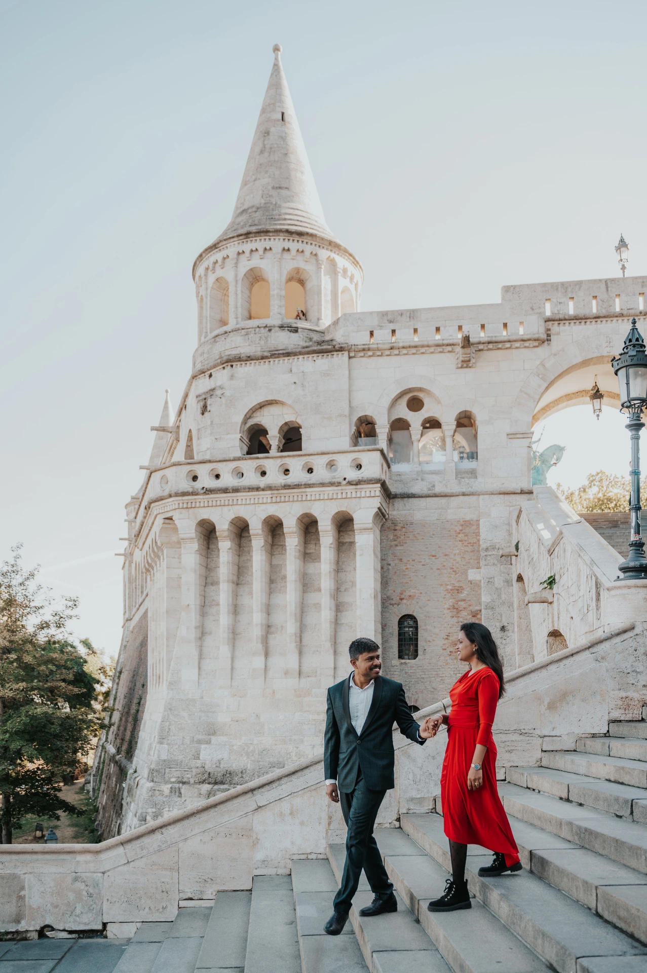 The couple walking hand in hand on a broad stone staircase with Fisherman’s Bastion’s tower and arcades behind them during their Budapest photoshoot.