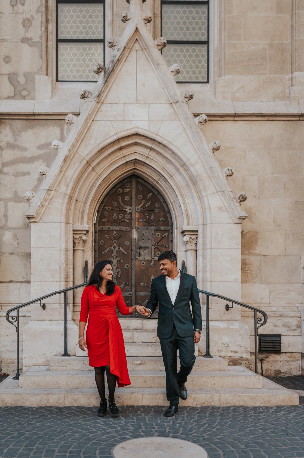The couple walking hand in hand down the stone steps in front of the ornate church doorway during their couple photoshoot in Budapest.