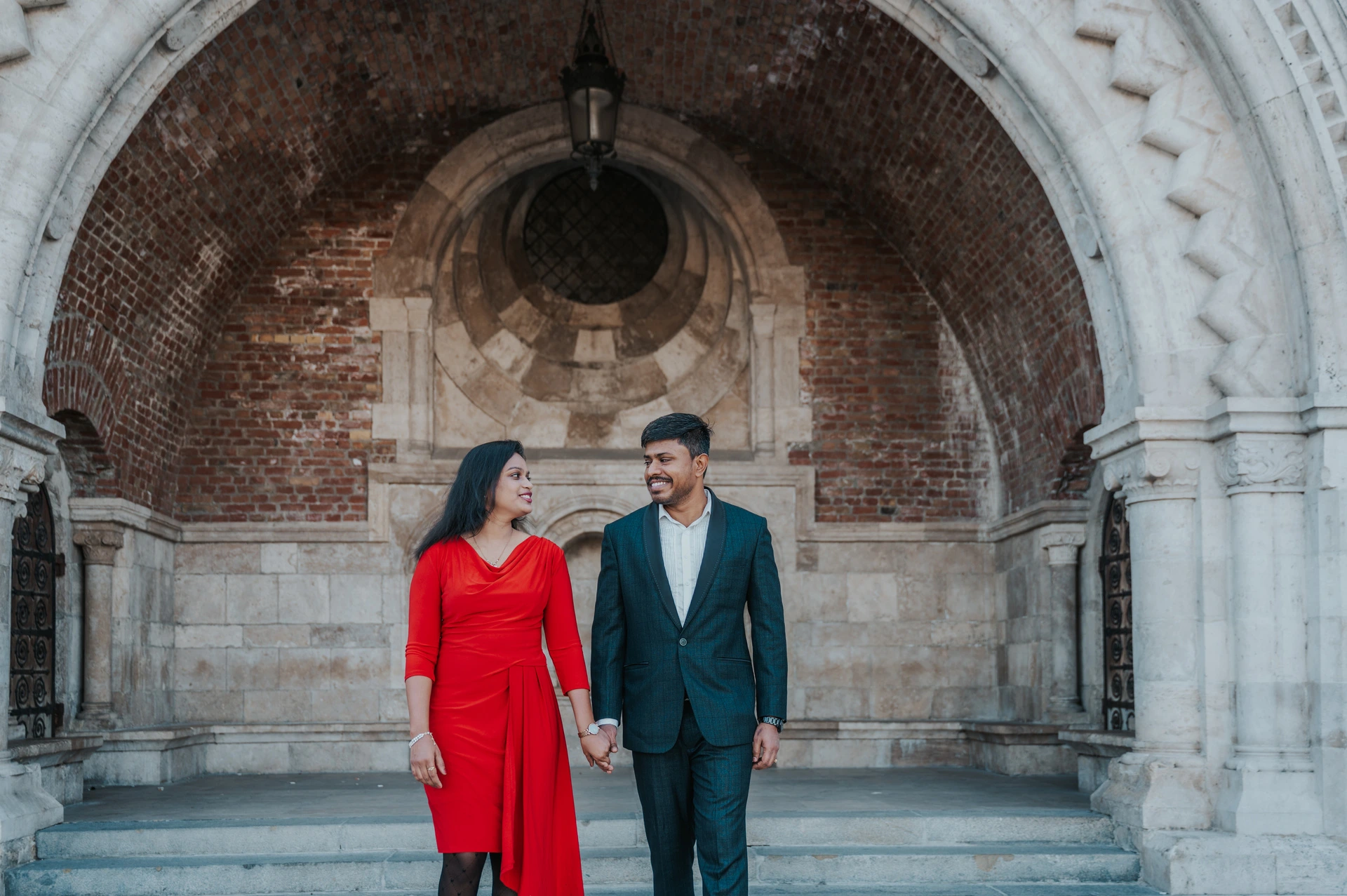 A wide landscape portrait of the couple holding hands beneath the brick-and-stone arch, with the hanging lantern above, during their Budapest photoshoot.
