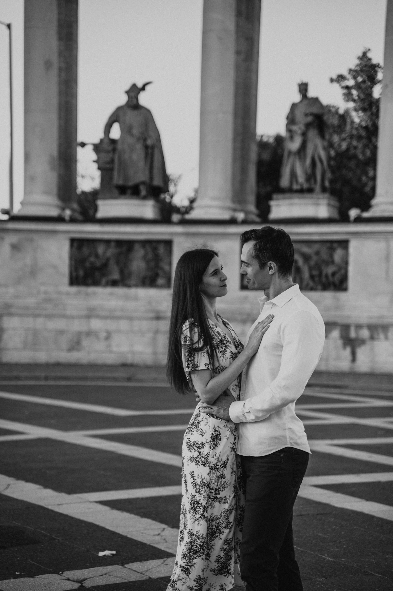 Black-and-white close portrait of the couple embracing at Heroes’ Square with bronze statues behind them during their couple photoshoot in Budapest.