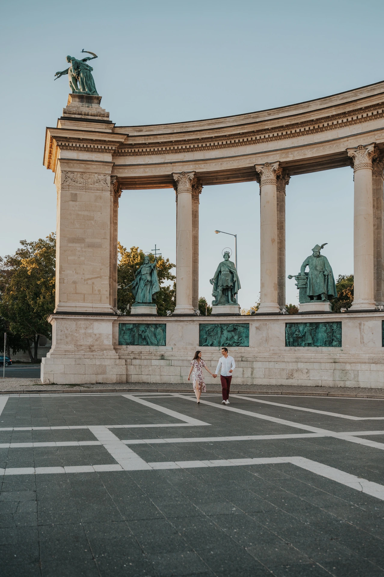 A wide portrait of the couple at Heroes’ Square with its stone columns, bronze statues and patterned paving during their couple photoshoot in Budapest.