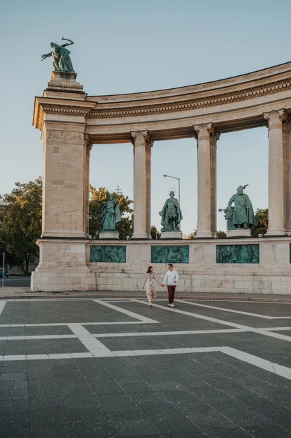 Columns and statues at Heroes’ Square at a Budapest couple photoshoot A wide portrait of the couple at Heroes’ Square with its stone columns, bronze statues and patterned paving during their couple photoshoot in Budapest.