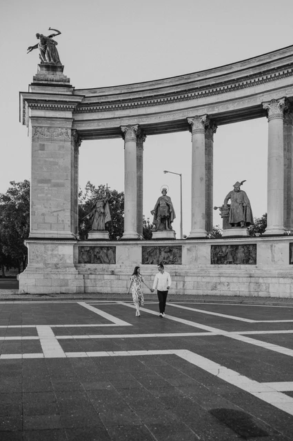 Black-and-white portrait of the couple walking across Heroes’ Square beneath its colonnade during their couple photoshoot in Budapest.