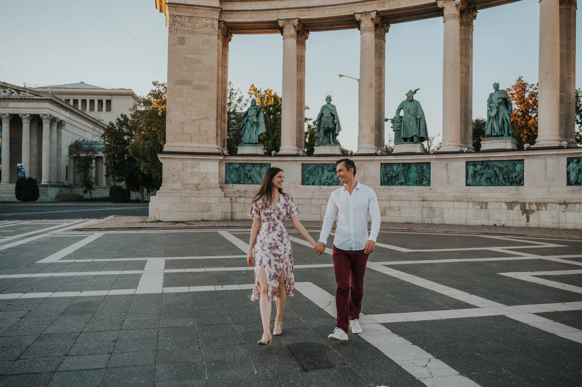 A landscape portrait of the couple walking across the patterned paving beneath Heroes’ Square’s colonnade during their couple photoshoot in Budapest.