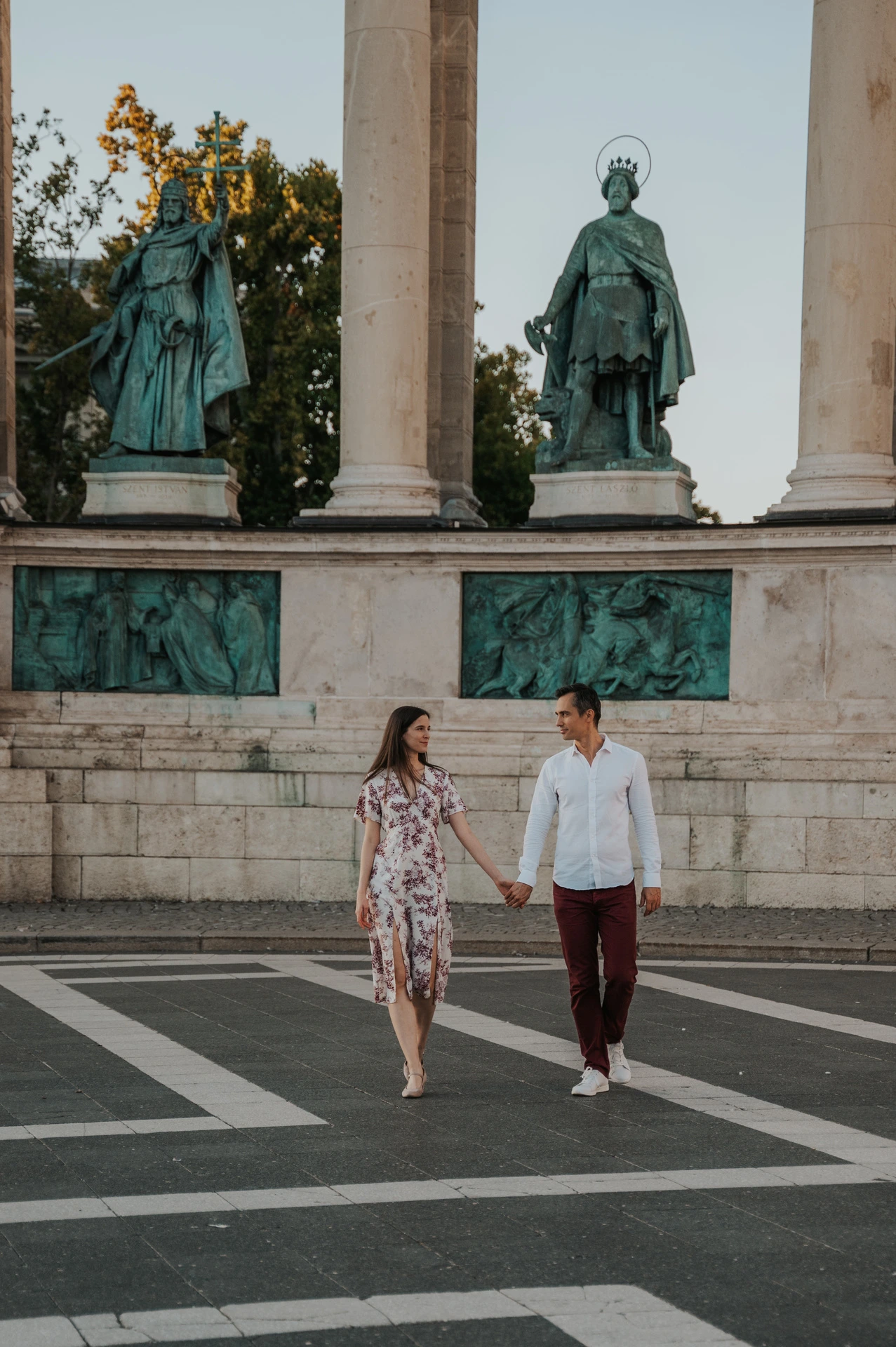 The couple walking together across Heroes’ Square with bronze statues and pale stone columns behind them during their couple photoshoot in Budapest.
