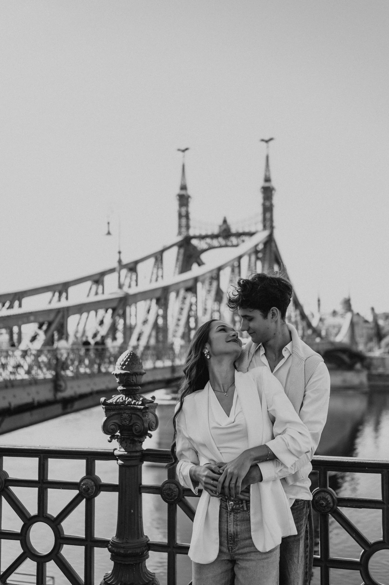 Black-and-white portrait of the couple embracing by the riverside railing during their Budapest photoshoot, with Liberty Bridge in the background.