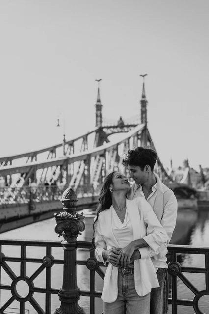 Budapest photoshoot with Liberty Bridge in black and white Black-and-white portrait of the couple embracing by the riverside railing during their Budapest photoshoot, with Liberty Bridge in the background.
