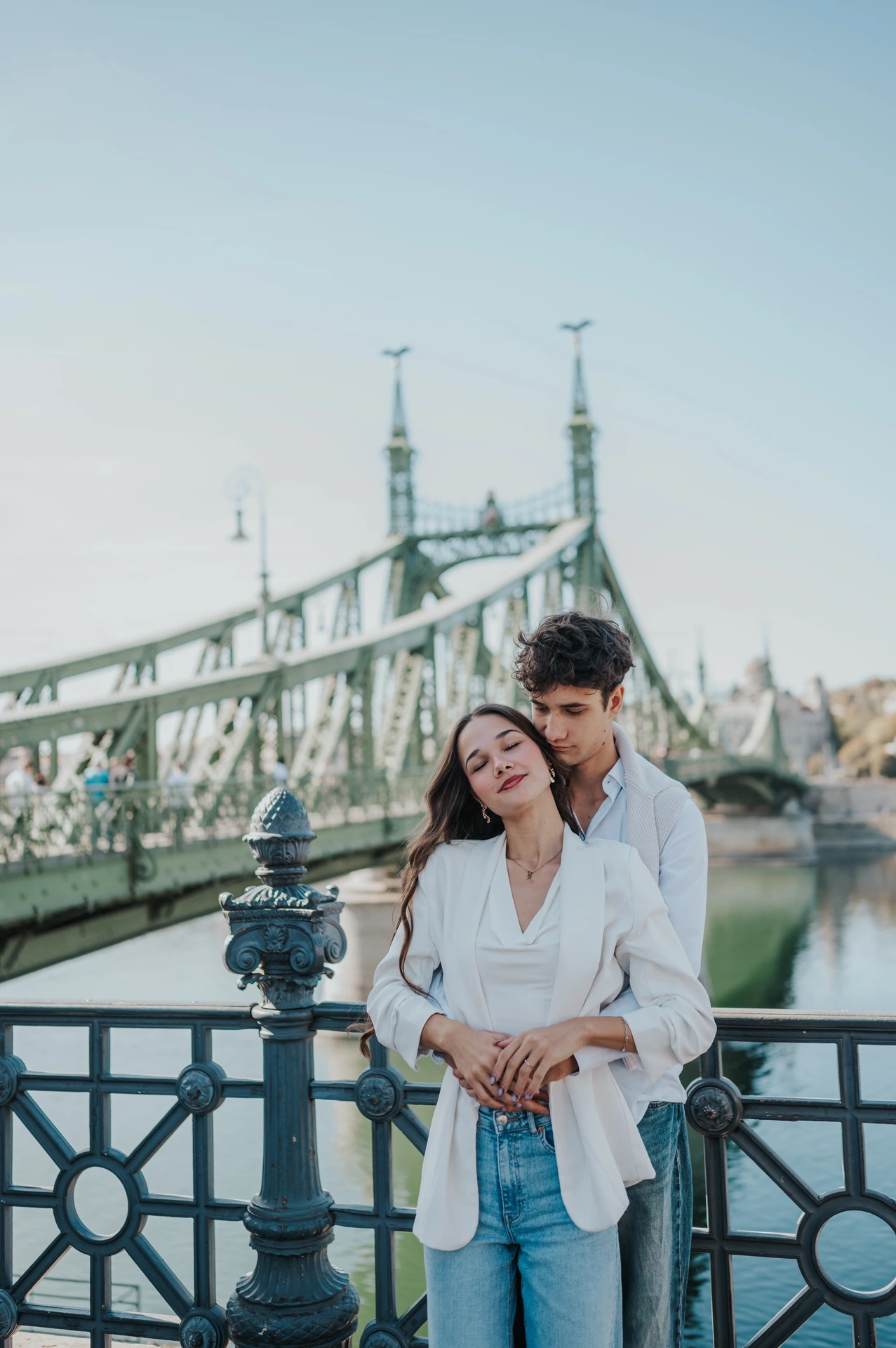 The man embracing his partner from behind during their Budapest photoshoot, with Liberty Bridge and the riverside railing around them.