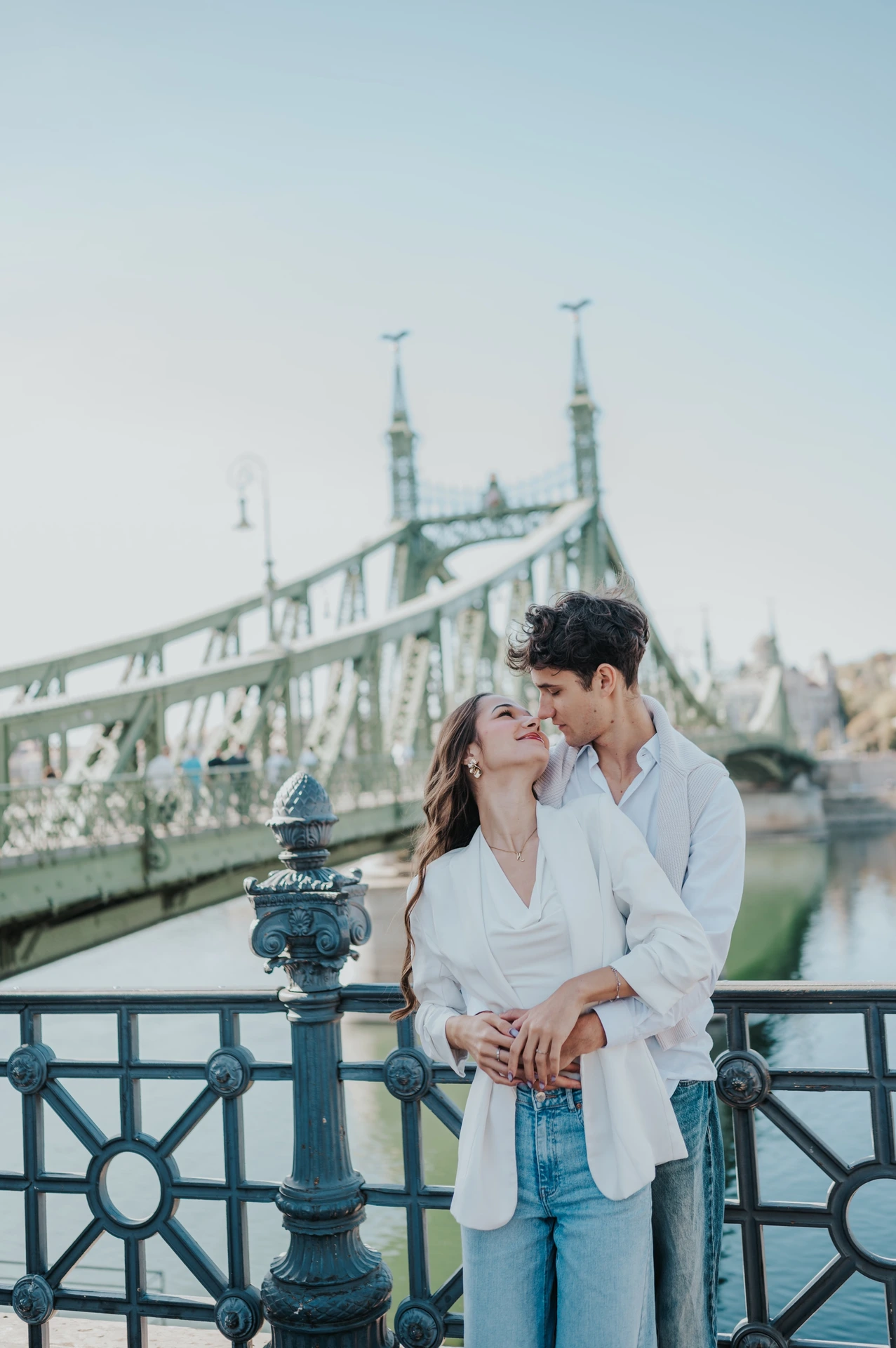 The couple leaning towards each other for a kiss beside the river during their Budapest photoshoot, with Liberty Bridge behind them.