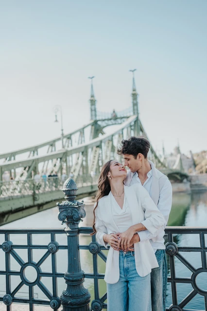 Budapest couple photoshoot sharing a kiss beside Liberty Bridge The couple leaning towards each other for a kiss beside the river during their Budapest photoshoot, with Liberty Bridge behind them.