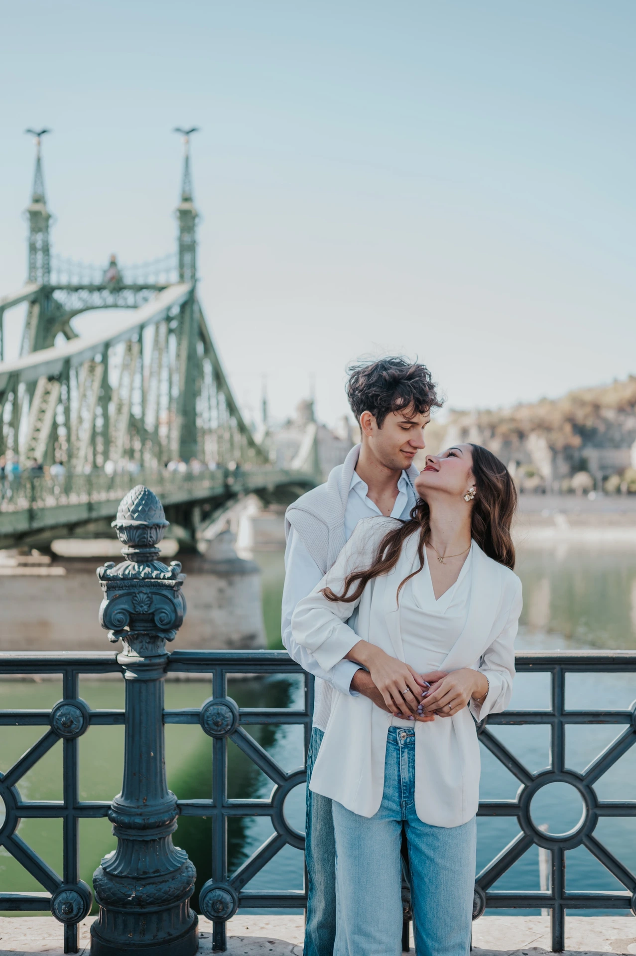 The couple holding each other close beside the decorative riverside railing during their Budapest photoshoot near Liberty Bridge.
