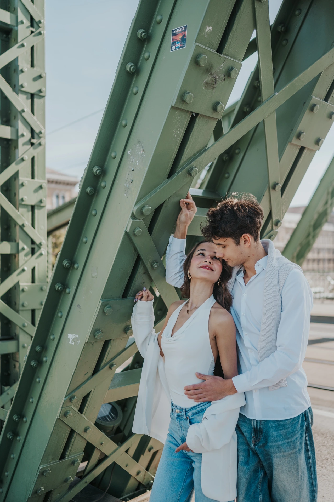 The man leaning towards his partner’s forehead as they embrace beneath the green beams during their Budapest photoshoot on Liberty Bridge.