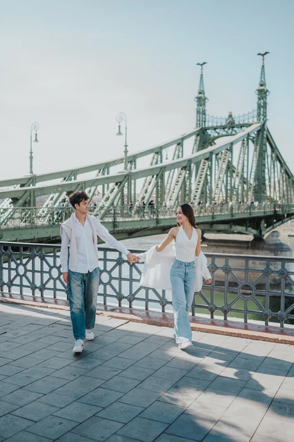The couple walking hand in hand beside the riverside railing during their Budapest photoshoot, with the green Liberty Bridge behind them.