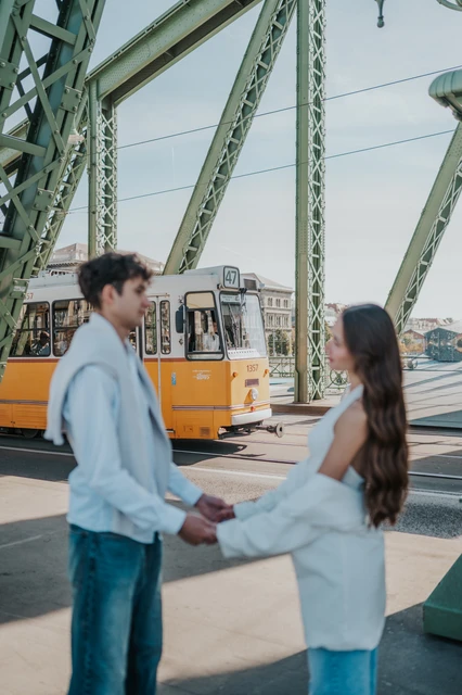 The couple facing each other and holding hands beneath the green steel beams during their Budapest photoshoot, with a yellow tram behind them.