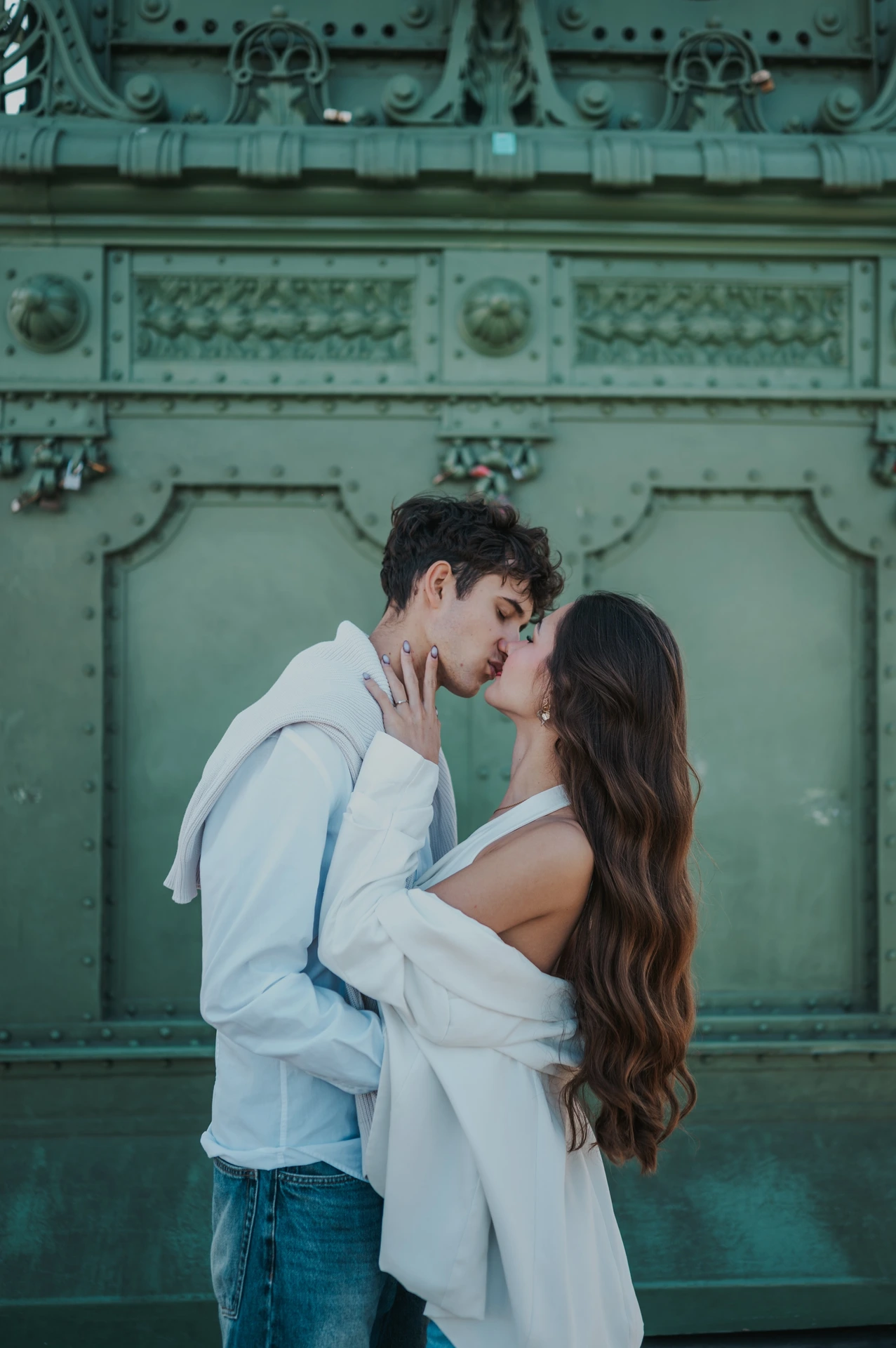 The couple kissing in front of the ornate green bridge pillar during their Budapest photoshoot, with her hand resting against his cheek.