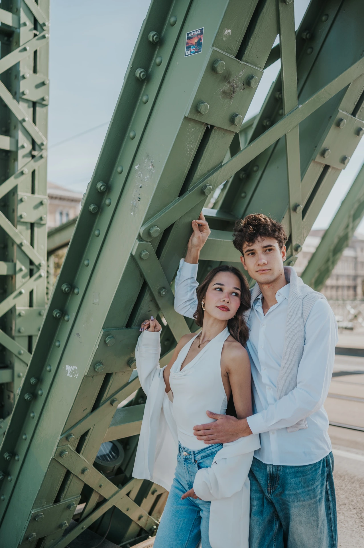 The couple smiling towards the camera beneath the green bridge structure during their Budapest photoshoot, with his arm raised above them.