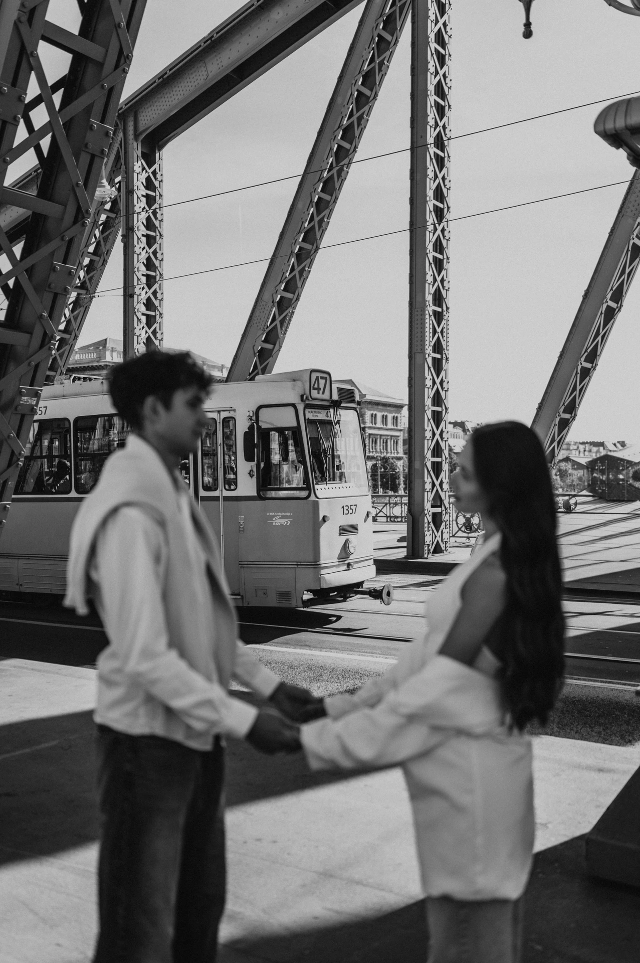 Black-and-white photograph of the couple facing each other and holding hands during their Budapest photoshoot, with a tram behind the Liberty Bridge beams.