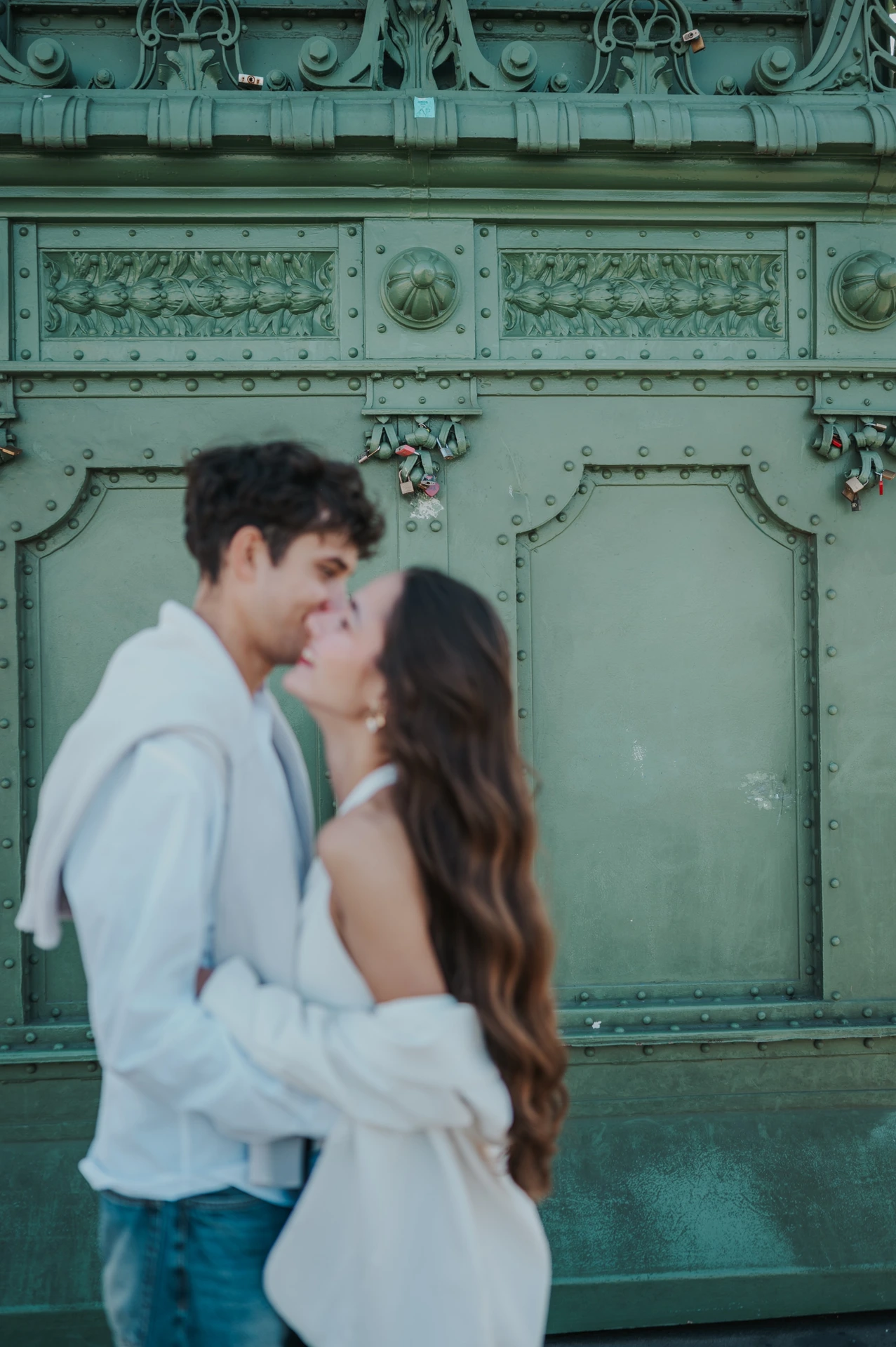 The couple holding each other close for a kiss during their Budapest photoshoot, framed against the riveted green ironwork of Liberty Bridge.