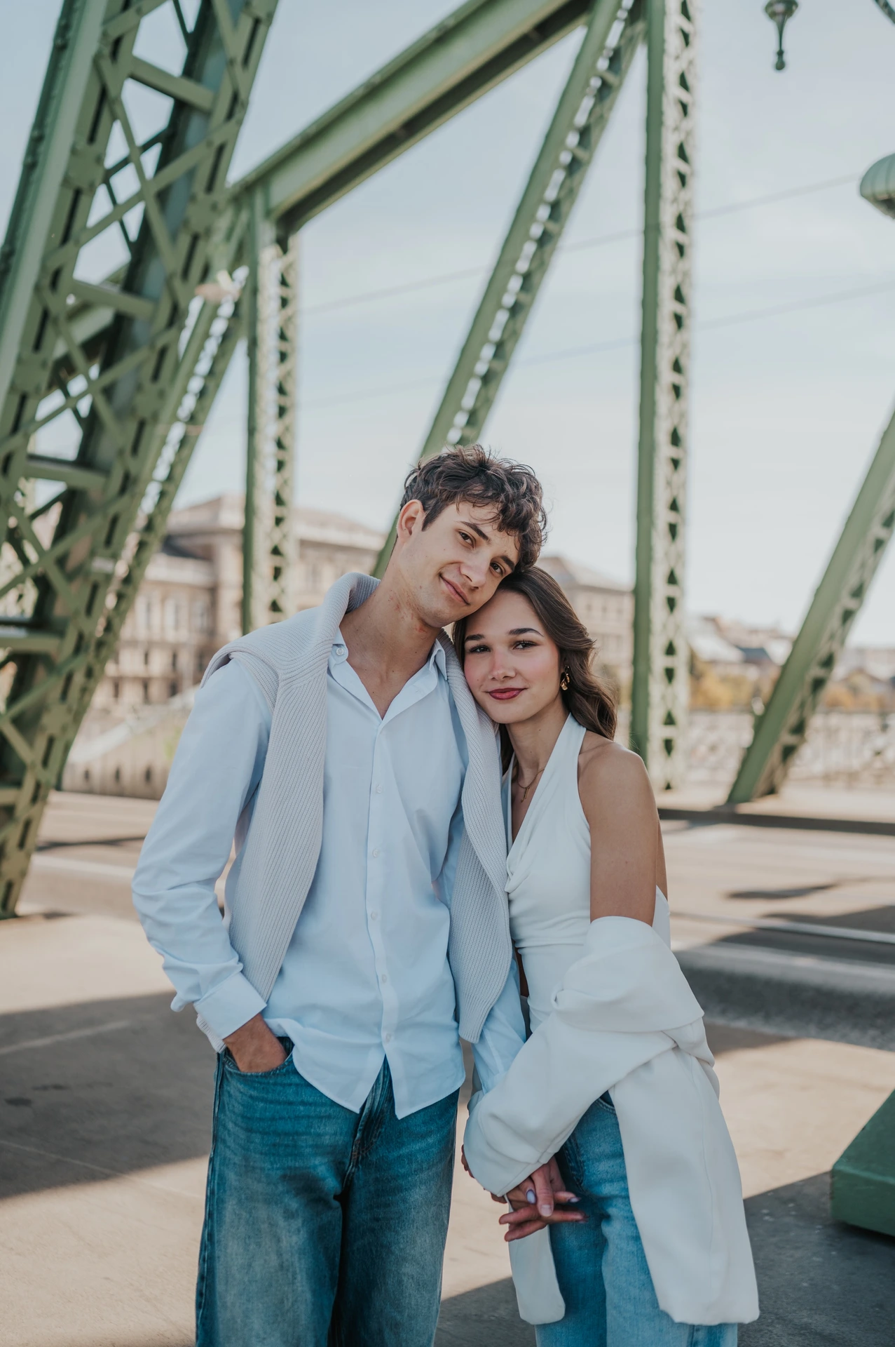 The couple leaning their heads together and looking towards the camera during their Budapest photoshoot beneath the green Liberty Bridge beams.