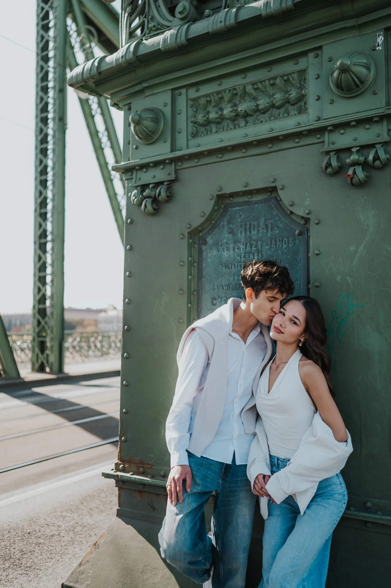 The woman resting her head beside her partner as they stand against the ornate green pillar during their Budapest photoshoot on Liberty Bridge.