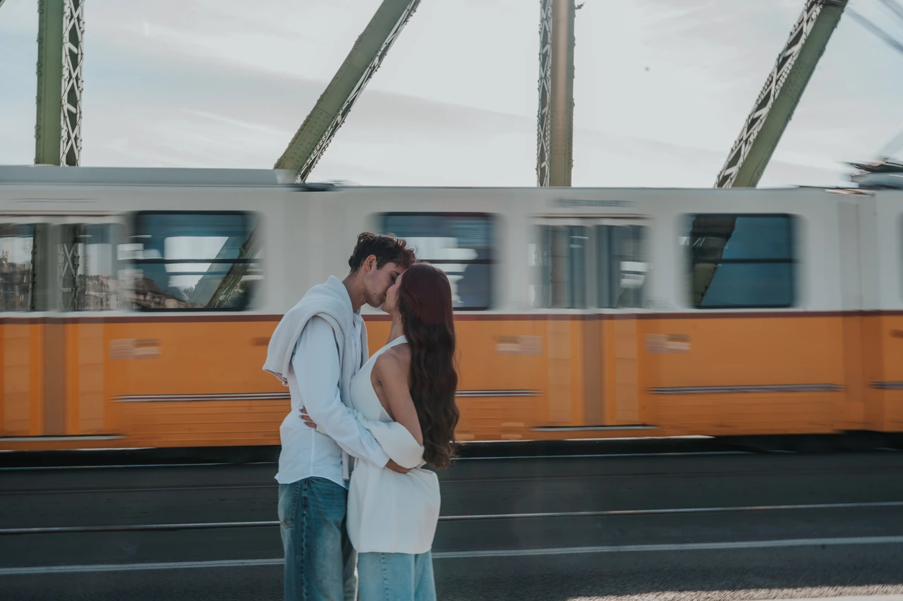 Photoshoot in Budapest with a passing tram on Liberty Bridge The couple kissing during their Budapest photoshoot on Liberty Bridge, with a yellow tram passing behind them and green beams overhead.