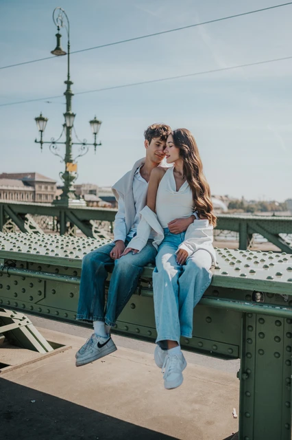 Budapest photoshoot seated beside a Liberty Bridge streetlamp The couple seated close together on the green railing during their Budapest photoshoot, with a decorative Liberty Bridge streetlamp behind them.