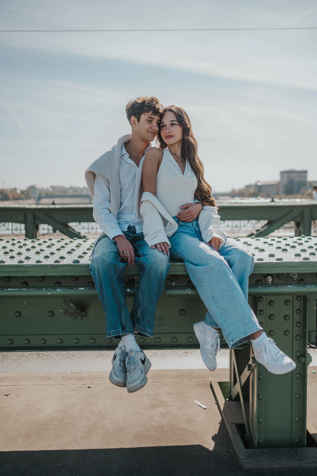 The man kissing his partner’s cheek while they sit on the bridge railing during their Budapest photoshoot, with her looking towards the camera.