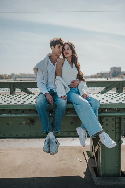 Budapest photoshoot sharing a cheek kiss on the Liberty Bridge railing The man kissing his partner’s cheek while they sit on the bridge railing during their Budapest photoshoot, with her looking towards the camera.