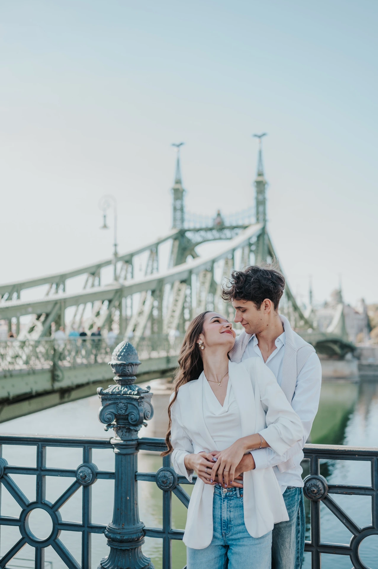 The couple looking towards each other beside the riverside railing during their Budapest photoshoot, with the green Liberty Bridge behind them.